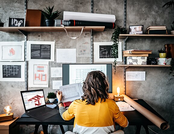 A woman sitting at a desk with printouts on the wall, a monitor, a Lenovo ThinkPad P15 Gen 2 mobile workstation open and showing architectural beams.